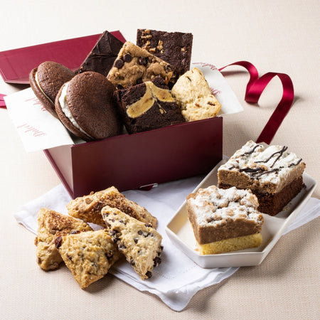 Assorted cookies and brownies in a gift box with a red ribbon on a beige surface.