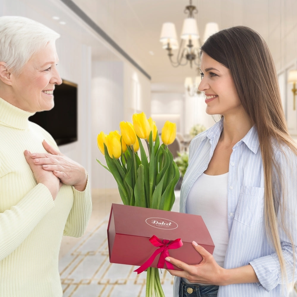 Woman holding a bouquet of yellow tulips and a red box with a pink ribbon, smiling at another person in a well-lit room.
